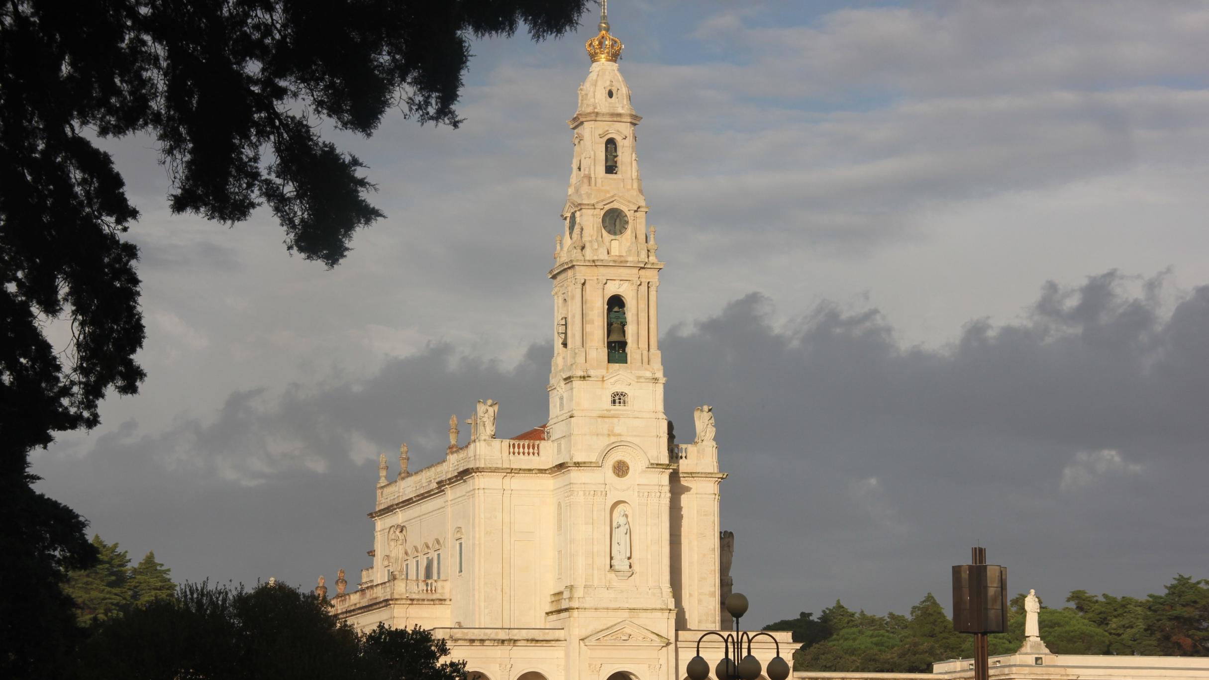 Sonnenbeschienene Basilika unserer lieben Frau des Rosenkranzes in Fátima unter wolkigem Himmel, Bäume im Vordergrund.