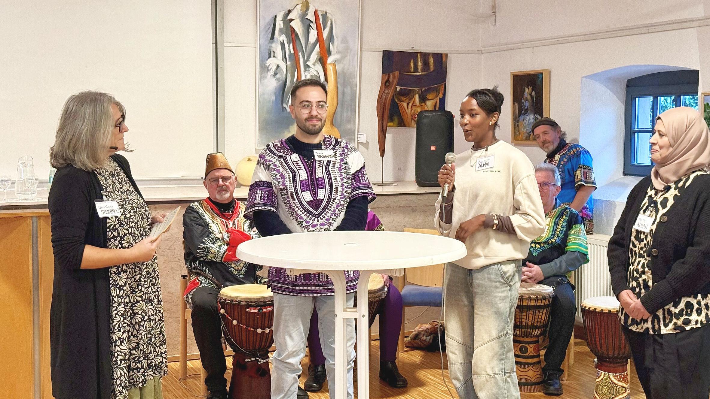 Vier Personen an einem Stehtisch, eine davon spricht in ein Mikrofon. Im Hintergrund sitzen Trommler in Tracht.