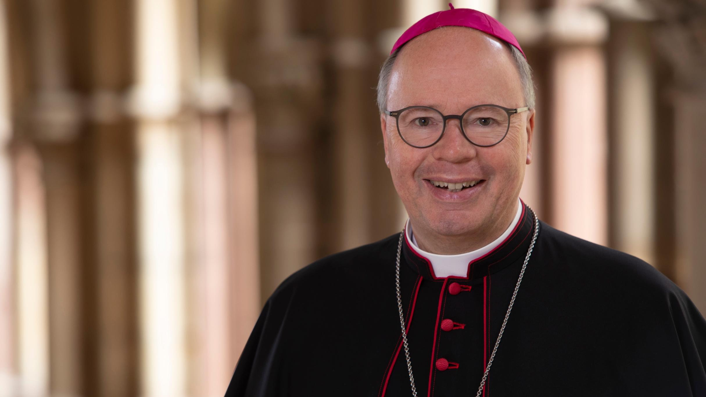 Hier ist ein Portrait von Bischof Ackermann, lächelnd im Kreuzgang zu sehen. Er Trägt ein Ornat.